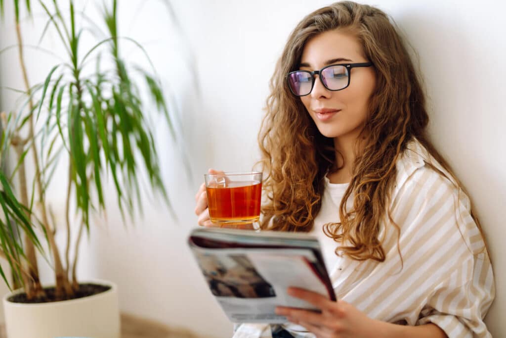 Young woman at home drinking coffee or tea and reading book or magazine